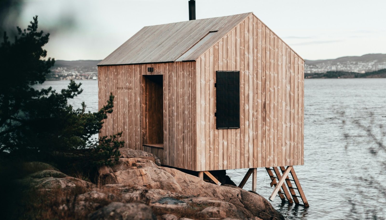 Holzhütte auf Stelzen am Wasser, umgeben von Bäumen und Bergen im Hintergrund.